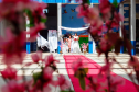 Imagem que mostra as noivas em fila preparadas para a entrada. O ambiente está decorado com panos brancos, flores rosas e um tapete vermelho.