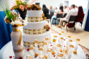 Imagem que mostra uma mesa decorada com um bolo, arranjo de flores e lembrancinhas em tons de branco e dourado. Ao fundo é possível ver os casais sentados, de costas.