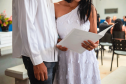 Imagem que mostra um dos casais segurando um documento. Ela usa um vestido branco com pequenas borboletas aplicadas, também brancas. Ele usa uma camisa branca e calça preta. A imagem não mostra o rosto deles.