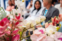 Imagem que mostra, em primeiro plano, flores de um dos arranjos da cerimônia. São orquídeas em tons de branco, rosa e vermelho. Ao fundo é possível ver um casal, de forma borrada.