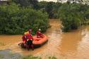 Imagem horizontal colorida em que se vê bombeiros em um bote inflável entrando em área alagada pelas enchentes no Litoral do Paraná. 
