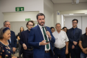 Defensor público-geral com um terno azul e gravata verde segura um microfone e um celular enquanto discursa para um público atento; ao seu redor, estão pessoas de diferentes idades e estilos, algumas vestindo roupas formais e outras mais casuais, em um ambiente fechado e iluminado.