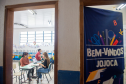 Imagem que mostra o interior de uma sala de aula vista através de uma porta entreaberta. Na sala, há pessoas sendo atendidas por agentes da DPE-PR. A sala possui janelas grandes com grades, permitindo a entrada de luz natural. Há decorações coloridas penduradas nas janelas e nas paredes. À direita, em primeiro plano, parte de uma faixa azul escura está visível, pendurada na porta, com a inscrição "BEM-VINDOS JOJOCA" em letras brancas grandes, acompanhada de desenhos coloridos de materiais escolares.