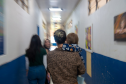 Imagem que mostra um corredor com paredes brancas na parte superior e azuis na parte inferior. Quatro pessoas estão caminhando no corredor, de costas para a câmera. A pessoa mais próxima carrega uma criança pequena em seus braços. As outras três pessoas são adultas, com cabelos longos e roupas casuais. As paredes do corredor estão decoradas com diversos quadros coloridos.