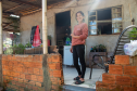 Imagem que mostra a senhora Amélia sorrindo em frente de sua casa. Ela tem cabelos escuros e veste uma blusa de manga comprida cor de telha, calças pretas e chinelos escuros. Ela está em uma área externa coberta, com piso de ladrilho claro. Ao fundo, à esquerda, há uma área mais aberta com vegetação e roupas penduradas em um varal improvisado. A casa é simples, tem paredes rebocadas e uma janela gradeada com cortinas brancas. A luz do sol entra pela lateral direita da imagem, criando um efeito de brilho.