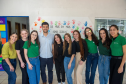 Imagem que mostra a equipe da Defensoria Pública. São nove pessoas, que sorriem posando para a foto em um ambiente interno. Há um homem e oito mulheres, todos com aparência jovem. A maioria das mulheres veste camisetas verdes e calças jeans. O homem veste uma camisa clara de botões e calças escuras. Ao fundo, na parede branca, há diversas impressões de mãos coloridas e a frase "DE MÃOS EM MÃOS SOMOS JOJOCAS" escrita em letras pretas. 