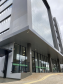 Imagem que mostra a nova sede da Defensoria em Foz do Iguaçu, com uma perspectiva mais ampla da fachada do prédio. O edifício apresenta um design moderno com múltiplas camadas e uma combinação de grandes painéis de vidro espelhados. Uma faixa horizontal verde clara com o logo da DPE-PR percorre a fachada. Uma rampa acessível com piso tátil amarelo leva à entrada principal e há também um corrimão de metal preto. O céu está nublado, o que confere uma iluminação difusa ao ambiente externo. 