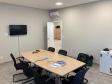 Imagem que mostra uma sala de reunião da nova sede da Defensoria em Foz do Iguaçu. Ela está equipada com uma mesa grande de madeira clara no centro. Ao redor da mesa, há várias cadeiras pretas e em cima da mesa, alguns cabos azuis de conexão à internet e documentos estão visíveis. Em uma das paredes, há uma televisão de tela plana montada e, à direita da imagem, um frigobar branco.
