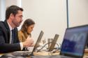 Um homem de terno e uma mulher acompanham reunião ao computador e em primeiro plano tela de laptop em que aparecem os participantes online da reunião. 