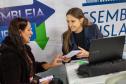 Imagem mostra duas mulheres sentadas, uma servidora ao computador entrega documentos a uma assistida.  