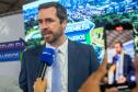 O DPG Matheus Munhoz dá entrevista. Homem de terno fala ao microfone, ao fundo tela de celular grava a entrevista. 