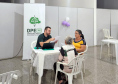 Imagem mostra duas mulheres, um idosa, sentadas à mesa conversando com homem jovem usando o computador, Ao fundo se vê banner da Defensoria com a logo da instituição.