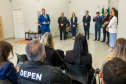 Nesta foto, o Defensor Público-Geral discursa de frente para a câmera. Em pé, logo atrás dele, um grupo de autoridades acompanha o pronunciamento. Em primeiro plano, vemos a plateia sentada de costas, assistindo à cerimônia. Um detalhe notável ao fundo, no canto esquerdo, é um pequeno espaço infantil com tapete colorido e brinquedos, sugerindo o caráter acolhedor do novo local.