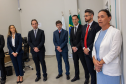 Em primeiro plano, no lado direito da imagem, a Ouvidora-Geral da DPE-PR faz seu discurso. Ela tem uma expressão séria e mantém as mãos unidas à frente do corpo. Ao seu lado, um grupo de autoridades, composto por quatro homens e uma mulher, ouve atentamente. Ao fundo, as bandeiras do Brasil e do Paraná estão hasteadas, compondo o cenário oficial.