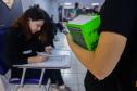 Imagem mostra braço de candidata segurando livro em close no primeiro plano enquanto fiscal confere seus documentos para ingressar na sala para do concurso. 
