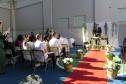 A imagem mostra a cerimônia de casamento em andamento. Vários casais estão sentados na primeira fila, com os homens vestindo camisas brancas. Ao fundo, convidados assistem. O pastor, de terno, fala ao microfone em um pequeno palco decorado com cortinas brancas e folhagens. Um tapete vermelho demarca o corredor central, ladeado por cestos com flores brancas. A luz do sol forte cria sombras pronunciadas.
