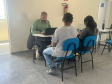 Imagem que mostra uma cena de atendimento individual. Três pessoas estão agrupadas em torno de uma pequena mesa branca. À esquerda, de frente para a mesa, está o atendente. Ele é um homem de pele morena, com cabelo escuro bem curto e barba. Veste uma camisa social de manga longa e está concentrado. Do outro lado da mesa, de costas para a câmera, estão duas pessoas sentadas em cadeiras com assentos e encostos azuis, um homem e uma mulher.