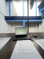 A imagem é uma visão em primeiro plano de uma mesa de trabalho de madeira. Sobre a mesa, há um laptop aberto e formulários de questionário para entrevistas. O ambiente é o pátio interno da cadeia, com paredes de concreto e estruturas pintadas de azul na parte superior.