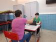 Em uma sala de aula, uma mulher de blusa rosa está sentada de costas para a câmera, em frente a uma mesa. Ela entrega um documento para uma servidora da Defensoria, uma jovem de camisa verde e óculos que está do outro lado da mesa, sorrindo e usando um laptop.