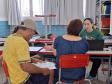 Dentro de uma sala de aula, um casal é atendido por uma servidora da Defensoria Pública de camisa verde. O homem, de camiseta amarela e boné, lê alguns papéis, enquanto a mulher, de blusa jeans e de costas para a foto, ouve a explicação da servidora.