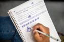 Imagem mostra mão com caneta e caderno em close onde se lê as anotações em letra cursiva Observatório de Violência contra  Mulher Indígena e desenhos com padrões guarani. 