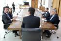 A foto é tirada por trás do governador, sentado na cabeceira de uma longa mesa de reuniões de madeira. De cada lado da mesa, duas pessoas em trajes sociais participam da conversa, sendo que um homem à esquerda gesticula enquanto fala. Ao fundo da sala ampla e formal, outras duas pessoas estão em pé.