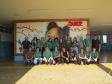 Uma foto de grupo com cerca de vinte pessoas, a equipe da Defensoria Pública, do CRAS Rural e da Escola Municipal posando em um pátio coberto de uma escola. Eles estão em pé e agachados em frente a um grande mural colorido pintado na parede ao fundo.