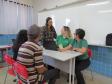 Em uma sala de aula, cinco pessoas estão reunidas ao redor de uma mesa branca com um laptop. Duas servidoras em camisas verdes e uma terceira mulher em pé atendem a um casal, que está sentado de costas para a câmera, em cadeiras vermelhas.