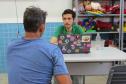 Dois homens conversam em uma mesa, um de frente para o outro. O homem de costas, com uma camisa azul, fala com um servidor mais jovem de camisa verde. Entre eles, sobre a mesa, há um laptop com vários adesivos. Ao fundo, prateleiras com brinquedos.