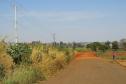 A imagem retrata uma paisagem rural com uma estrada de terra vermelha que se estende ao horizonte. A lateral da estrada é coberta por vegetação alta e seca, e há postes de energia elétrica. Ao fundo, uma extensa área de campo e árvores sob um céu claro.