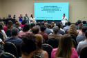 Um auditório cheio com a plateia sentada de costas para a foto, voltada para um palco. No palco, uma mulher de branco fala ao microfone ao lado de uma tela projetando o título de um curso sobre heteroidentificação racial.