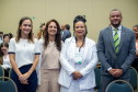 Quatro pessoas posam lado a lado para uma foto em um ambiente interno. Da esquerda para a direita: uma mulher de blusa branca, uma mulher de roupa bege, uma mulher negra com tranças e roupa branca, e um homem negro de terno cinza e gravata verde.