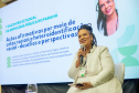 Uma mulher negra com tranças e roupa branca sorri enquanto fala ao microfone, sentada no palco. Ao fundo, uma tela projeta sua foto, nome e o tema da palestra sobre ações afirmativas e cotas raciais.