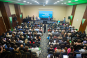 Vista superior do fundo de um auditório lotado, com a plateia voltada para um palco com iluminação azul. No telão, lê-se "Modelos de Reparação Coletiva da DPMG", enquanto palestrantes debatem sentados no palco.