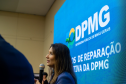 Uma mulher de perfil, com cabelos longos, fala ao microfone. Atrás dela, um painel de LED azul exibe o logotipo da DPMG e o texto parcial "Modelos de Reparação Coletiva".