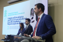 Um homem de terno azul e gravata vermelha discursa em um púlpito. Atrás dele, duas pessoas estão sentadas no palco, e um telão exibe o título da palestra: "Litigância Estratégica com foco no extrajudicial".