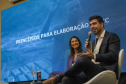 Um homem de terno escuro e camisa branca fala ao microfone, sentado em uma poltrona branca no palco. Ao seu lado, uma mulher de blazer azul sorri enquanto observa a plateia. Atrás deles, um grande painel de LED azul exibe a frase "PRINCÍPIOS PARA ELABORAÇÃO".