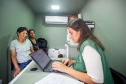 Defensora Mariana, com colete verde da Defensoria, atende duas mulheres, uma com blusa azul claro e outra com camiseta escura. Elas estão no interior da van dos direitos. Ao fundo um ar condicionado e uma mesinha com impressora. 
