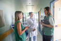 Defensora Ingrid e defensora Mariana, conversando com funcionária de hospital local. Elas estão em um corredor com paredes claras, ao fundo uma luz de contraste com funcionária. 