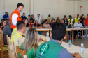 Em uma sala de reunião, o secretário das cidades do Estado, Guto Silva, está em pé vestindo colete laranja (Defesa Civil/Casa Militar), fala com o público sentado nas mesas brancas. Em primeiro plano, uma mulher e um homem de colete verde da Defensoria Pública ouvem.