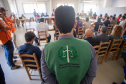 Cena de reunião com várias pessoas sentadas em mesas brancas em "U" em uma sala iluminada. Em primeiro plano, defensores públicos juntos de oficiais da Polícia Militar e outras autoridades, sentados. Há uma tela de projeção exibindo um mapa ao fundo.