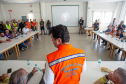 Em uma sala de reunião, o secretário das cidades do Estado, Guto Silva, está em pé vestindo colete laranja (Defesa Civil/Casa Militar), fala com o público sentado nas mesas brancas.