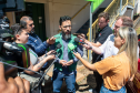 O defensor público, Fernando Redede - um homem de barba e colete verde da Defensoria Pública - é cercado por repórteres que seguram microfones com logotipos de emissoras, em uma entrevista rápida ao ar livre.