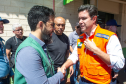 Dois homens brancos se cumprimentam com um aperto de mãos. O da esquerda, o defensor público Fernando Redede, com barba e óculos, veste um colete verde da defensoria. À direita, o secretário das cidades do Estado, Guto Silva, veste um colete de segurança laranja, com a mão esquerda sobre o peito. Há outras pessoas ao fundo.