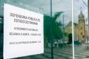 Imagem da placa da Defensoria com reflexo na porta de vidro da Câmara em que se vê o reflexo da igreja na praça. da igreja da praça em Matinhos. 
