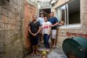 Família com quatro pessoas adultas posa em frente à sua casa. Muros e paredes sem reboco compõe o cenário. 