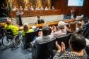 Cadeirante em primeiro plano e público ao redor aplaudem lançamento do NUPED na ALEP.  