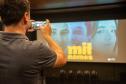 Um homem de costas fotografa com celular a tela com cartaz do documentário Mil Nomes.