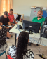 Uma sala com um funcionário de camisa verde atendendo em um computador. À frente dele, um grupo de pessoas aguarda sentado.