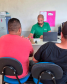 Um close do atendimento onde o funcionário de camisa verde consulta documentos e um notebook. De frente para ele, dois homens aguardam sentados de costas para a câmera em cadeiras de assento azul.